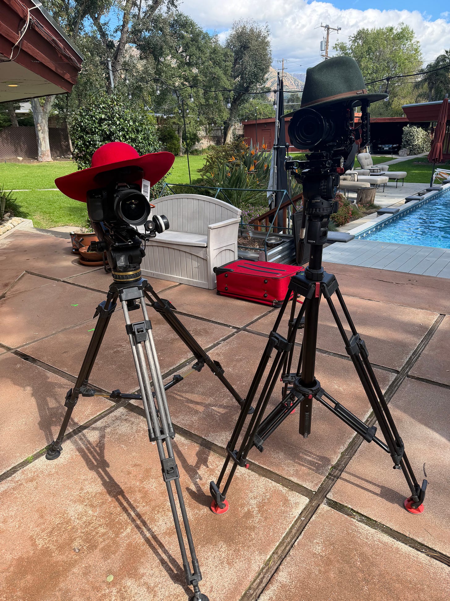 Two cinema cameras on tripods sit on a backyard patio beside a pool during a shoot. One camera is topped with a bright red wide-brim hat, the other with a green felt hat. A red equipment case rests nearby, with outdoor seating, garden plants, and a mid-century house in the background under a partly cloudy sky.