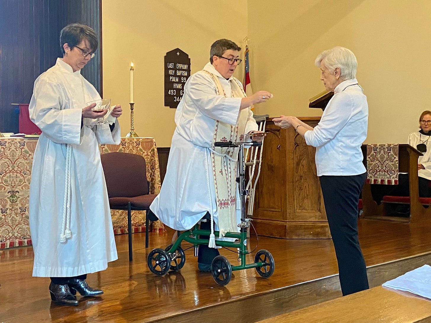 Woman receiving communion from female priest, with another woman in a robe assisting.