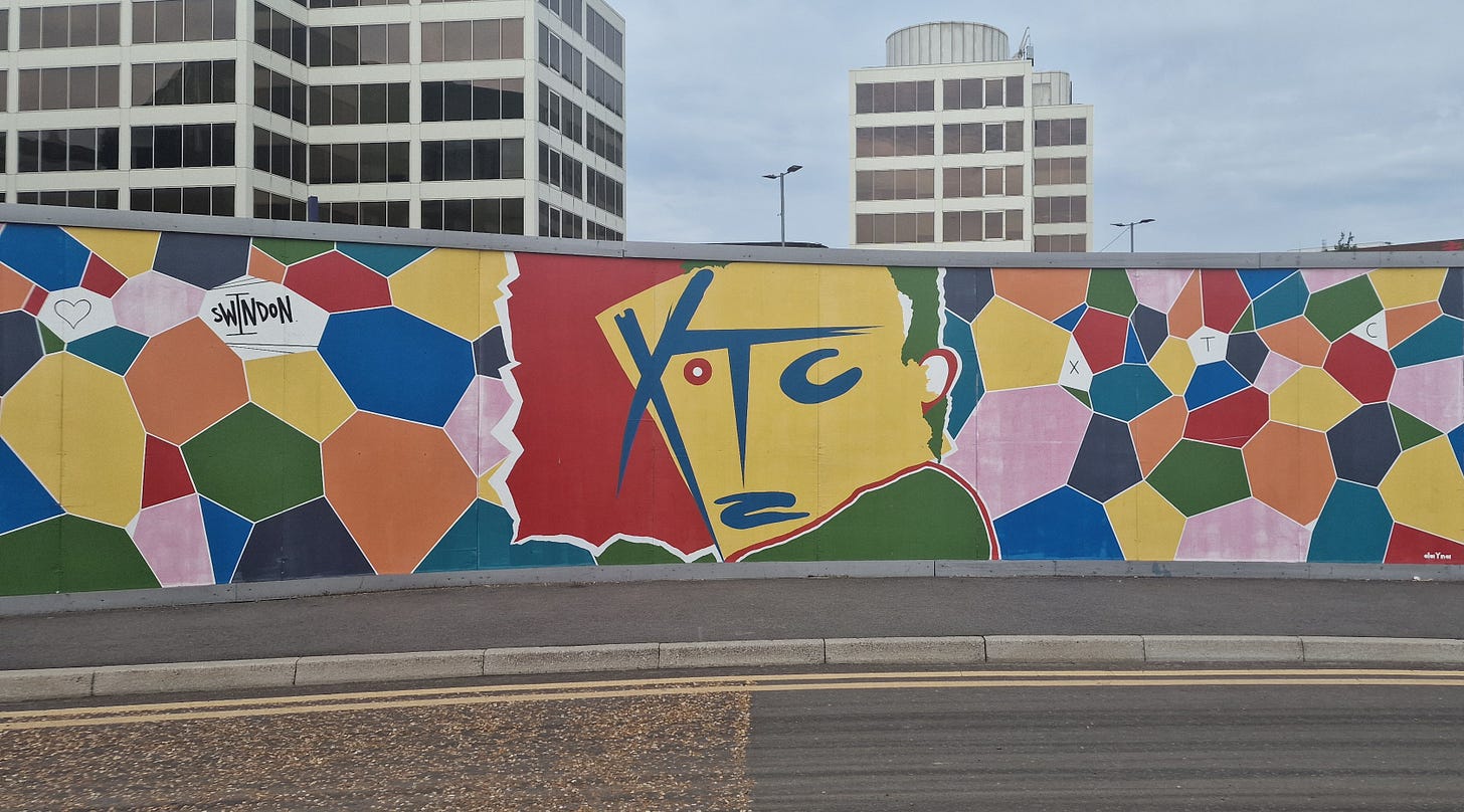 A stretch of wall with a couple of tall buildings in the background. On the wall are colourful hexagonal shapes and in the middle is a replica of the colourful image from XTC's Drums and Wires album.