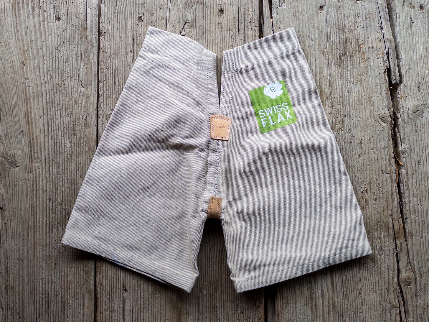 Kurze, naturfarbene Schwingerhose aus robustem Schweizer Leinen (Swiss Flax) mit Lederverstärkungen an der Mittelnaht; oben rechts ist ein grünes Logo mit weisser Flachsblüte und dem Schriftzug „SWISS FLAX“ aufgedruckt. Die Hose liegt flach ausgebreitet auf verwitterten Holzbrettern. Kurze, naturfarbene Schwingerhose aus robustem Schweizer Leinen (Swiss Flax) mit Lederverstärkungen an der Mittelnaht; oben rechts ist ein grünes Logo mit weisser Flachsblüte und dem Schriftzug „SWISS FLAX“ aufgedruckt. Die Hose liegt flach ausgebreitet auf verwitterten Holzbrettern.