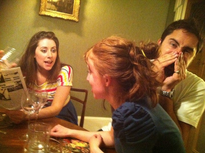 Three people at a table. Two women are mid animated conversation and a man is leaning into the camera pulling a weird face 