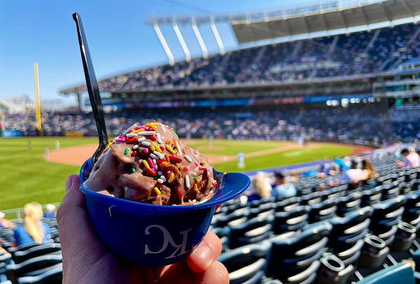 Chocolate peanut butter ice cream with rainbow sprinkles in a blue Kansas City Royals helmet cup. The picture is taken from the third base side of the field looking at home plate, with the right field foul pole in the distance in the top left corner of the picture.