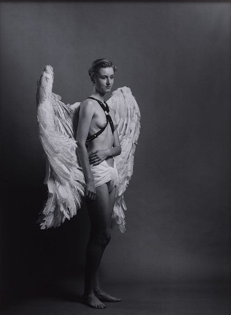 1) A black-and-white photo of a white woman with short dark hair, wearing large white feathered angel wings, a leather chest harness, and white shorts. The tone is dramatic and ethereal. She is standing to her side, with her left arm holding her stomach, and she is looking down and away from the camera. Her chest is bare, with the thin leather harness over it. 2) A woman in historical attire poses confidently in a black and white photo. They hold a feathered mask on a stick, wearing a striped coat and breeches. They have on traditional colonial shoes, and their white undershirt is long-sleeved with ruffled wrists. Her front left leg is out in front and turned to the side, while her chest is stuck out, and her left arm at her side, while the right arm holds the mask stick. She has short dark hair. 3) A woman in 18th-century attire poses elegantly in a black-and-white photograph. She wears a voluminous Victorian gown and a white powdered wig, holding a beaded mask in one hand and a sign in the other. The sign on her right reads: 'Una Troubridge, Alice Austen, Janet Flanner, Sappho, Radclyffe Hall'.