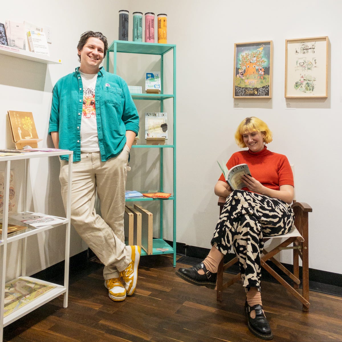 Two people are in a brightly lit gallery-style bookshop corner, surrounded by zines and small press publications. The person on the left stands casually with one hand in a pocket, wearing a teal button-up shirt over a graphic T-shirt, beige pants, and yellow-and-white New Balance sneakers. The other, seated in a wooden director's chair on the right, has short blonde hair and is wearing a red short-sleeve turtleneck with black and white abstract-patterned pants, reading a book with a smile. Behind them are teal and white shelving units displaying colorful books and zines, and the wall features framed art prints in an illustration style.