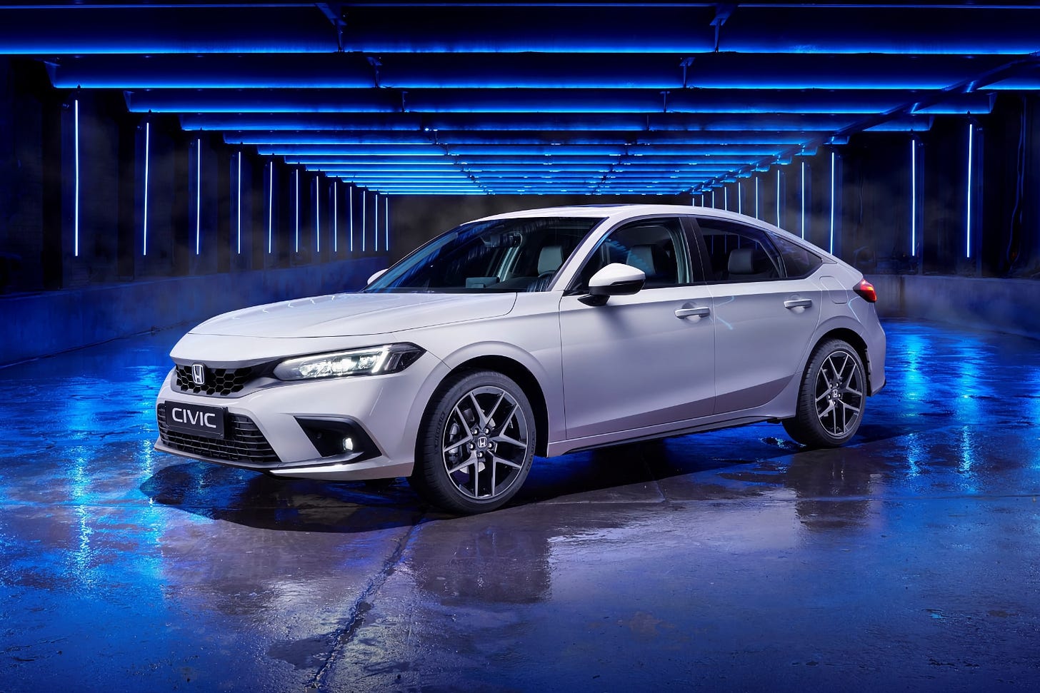 A white Honda Civic Hybrid hatchback parked on wet pavement surrounded by blue lighting at night.