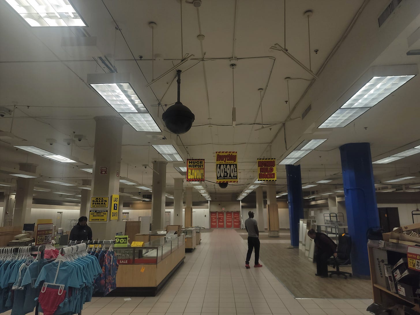 A nearly empty store. To the left, a rack full of clothes. A single customer stares into the void beyond. A woman sits on a chair next to a blue column. The ceiling is bare. Everything is very sad