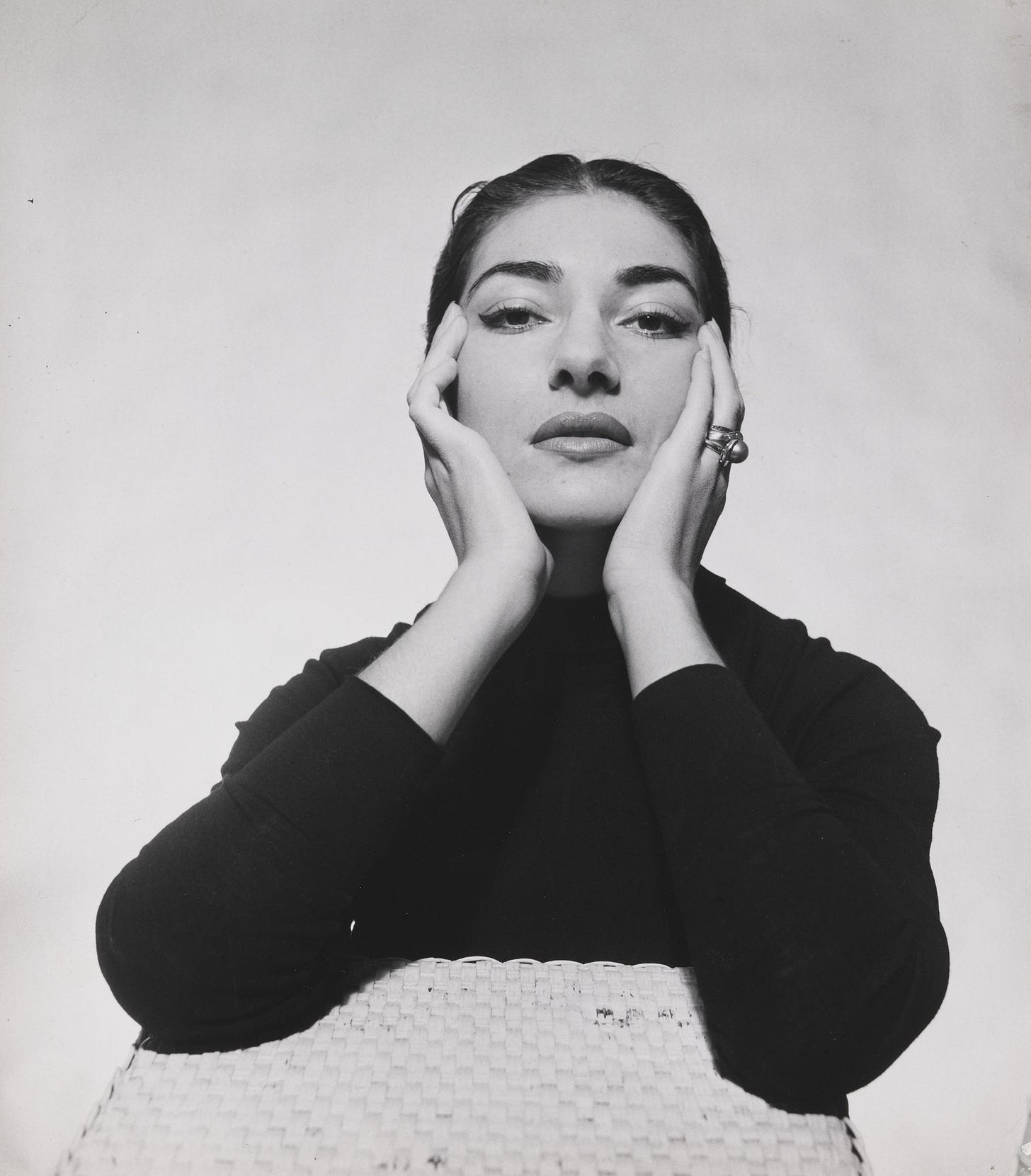 A black and white closeup portrait of a dark haried woman wearing a black sweater as she gazed directly into the camera lens while cradling her face in her hands.
