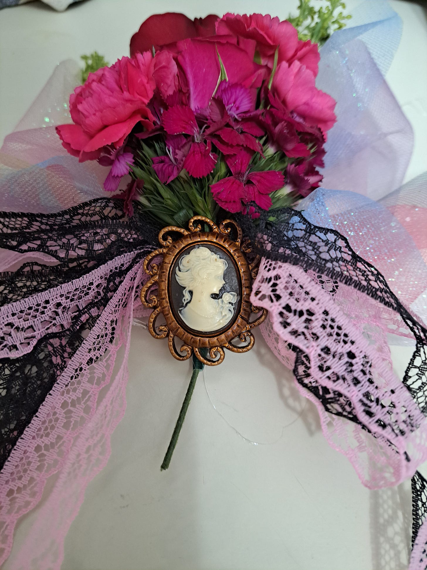A corsage of dark pink, purple, and red flowers, with a glittery background and a pink and black bow. In the bow is a cameo.