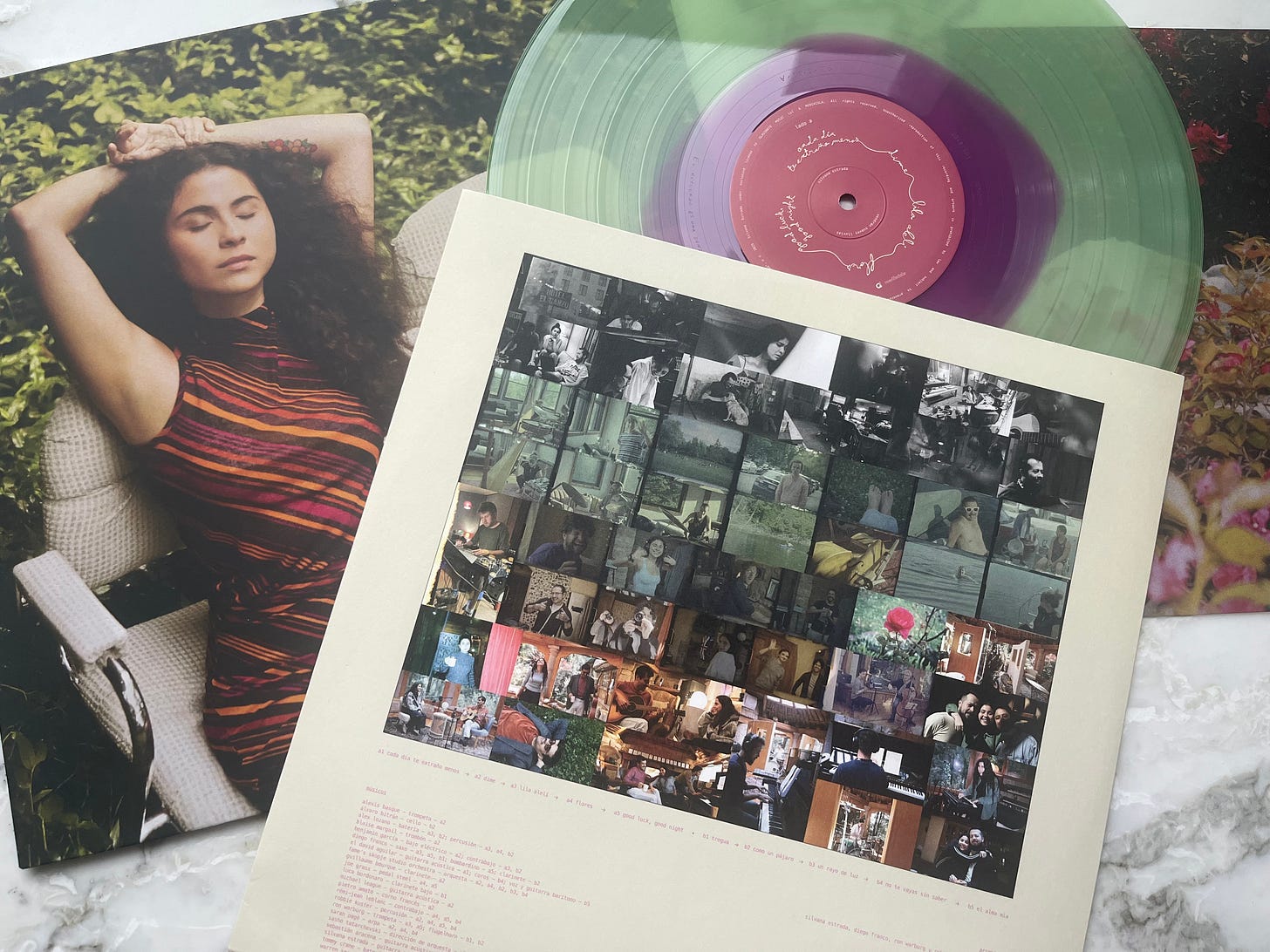 A semi-translucent green and purple LP record, half out of its sleeve that shows a grid or many photographs, beside the outer album cover with a photo of Silvana Estrada reclining in a lawn chair