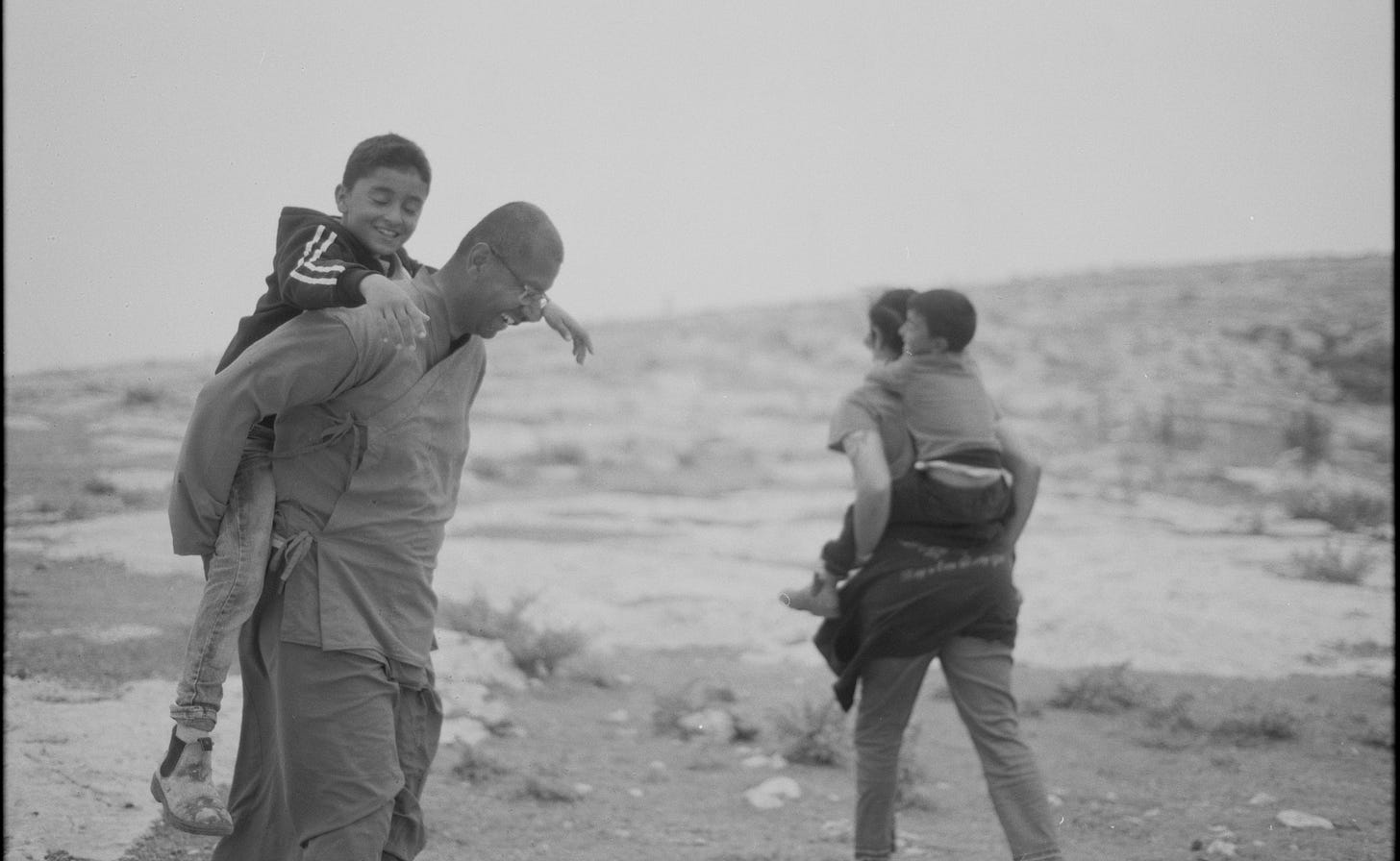 A black and white photo of two Palestinian children on two different adult’s shoulders, including a Buddhist monk.