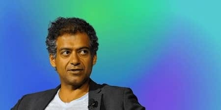 A man with curly dark hair and wearing a gray blazer, appearing thoughtful while speaking at an event with a colorful gradient background. A man with curly dark hair and wearing a gray blazer, appearing thoughtful while speaking at an event with a colorful gradient background.