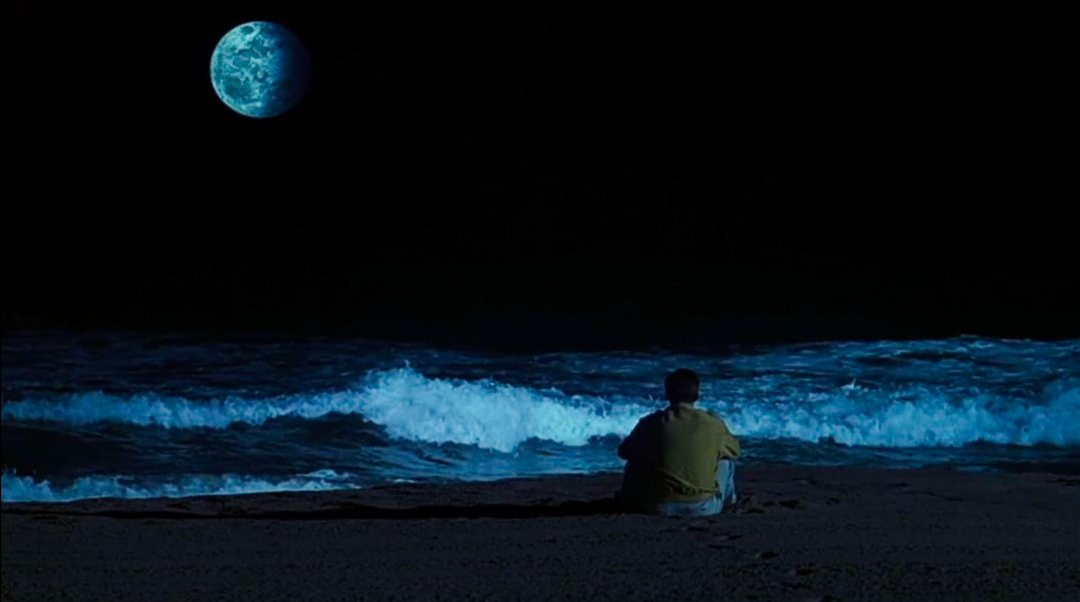 A lone man sits on a dark beach at night, facing crashing waves beneath an artificial-looking moon in the sky. | The Truman Show (1998)
