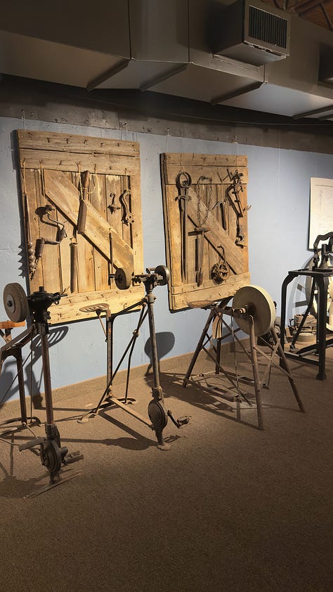 A covered wagon and a Pioneer cabin in spotlight on the lower level of a museum. | Displays of Pioneer tools. | A girl with two Dutch braids sits at a table with plaid cloth.