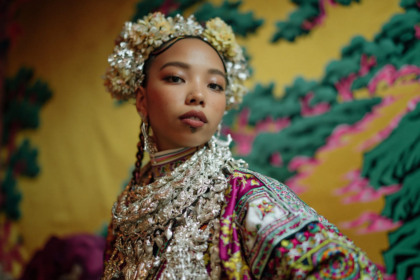 Candid photograph of a young Vietnamese Hmong woman wearing traditional jewelry, clothing, and hair styling; heavy, layered silver necklaces and intricate floral hairpieces. Her body is half-turned away, and she is looking back at the camera as if surprised. The background is a traditional Vietnamese silk painting showing a river and trees, in yellow and green with pink highlights. Dutch angle, three-quarter portrait.