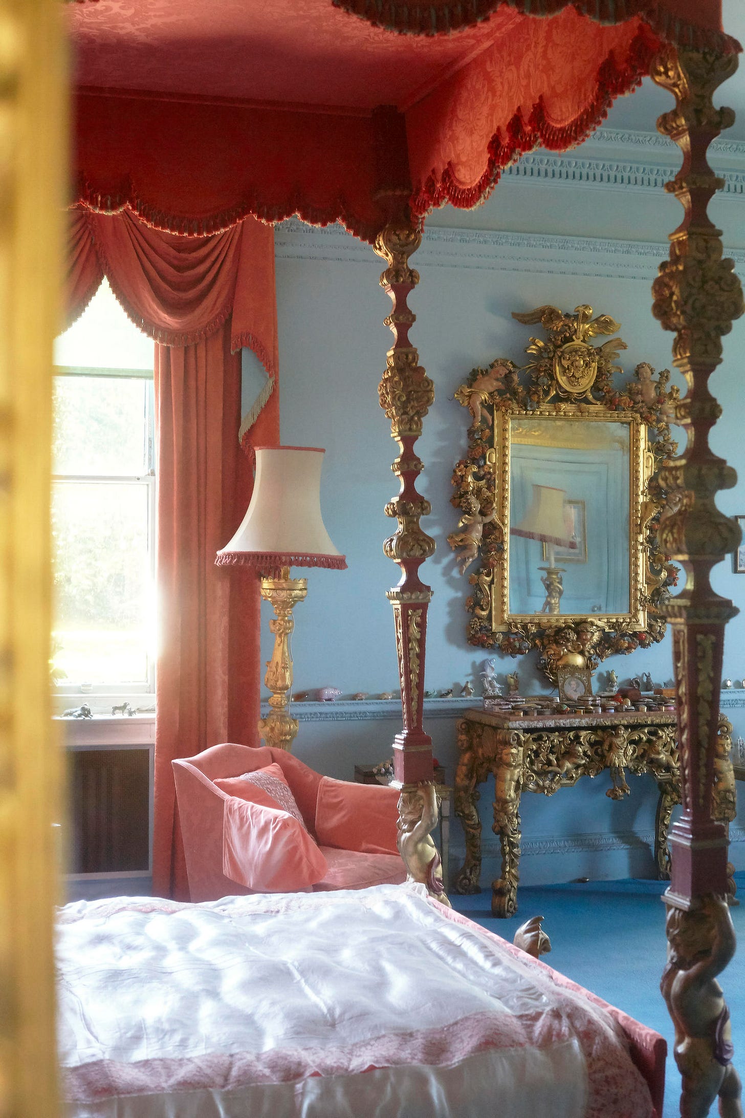 Elegant bedroom with ornate gold mirror and table. Red canopy bed, matching curtains, and lamp. Calm mood with soft light filtering in. Dame Barbra Cartland's Bedroom Photographed By Claudia Legge