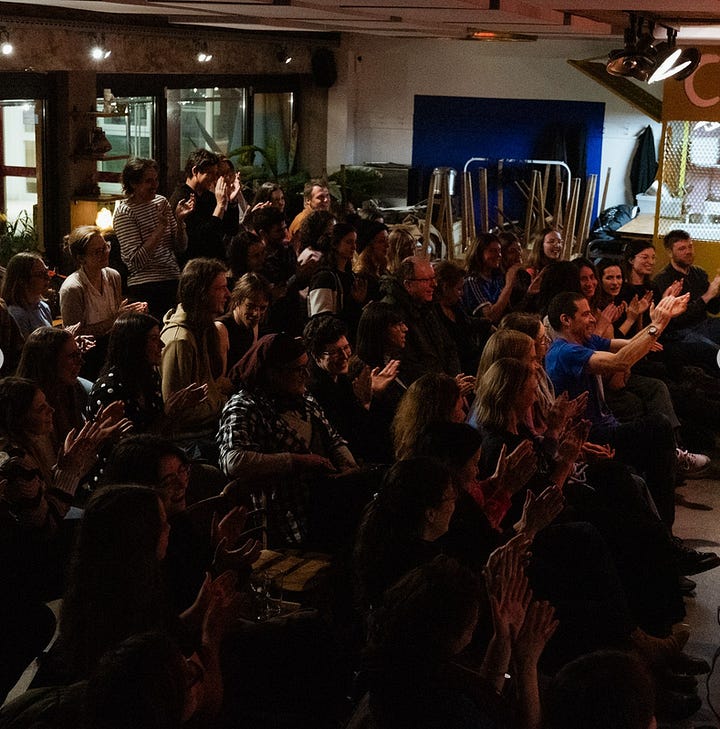 In the left-hand photo, a packed audience watches a performer off-camera in a low-lit bar in the evening. They are applauding. In the right-hand photo, a man sits on a stool on a stage with his back turned to the camera speaking into a mic to a listening audience. The audience are laughing.