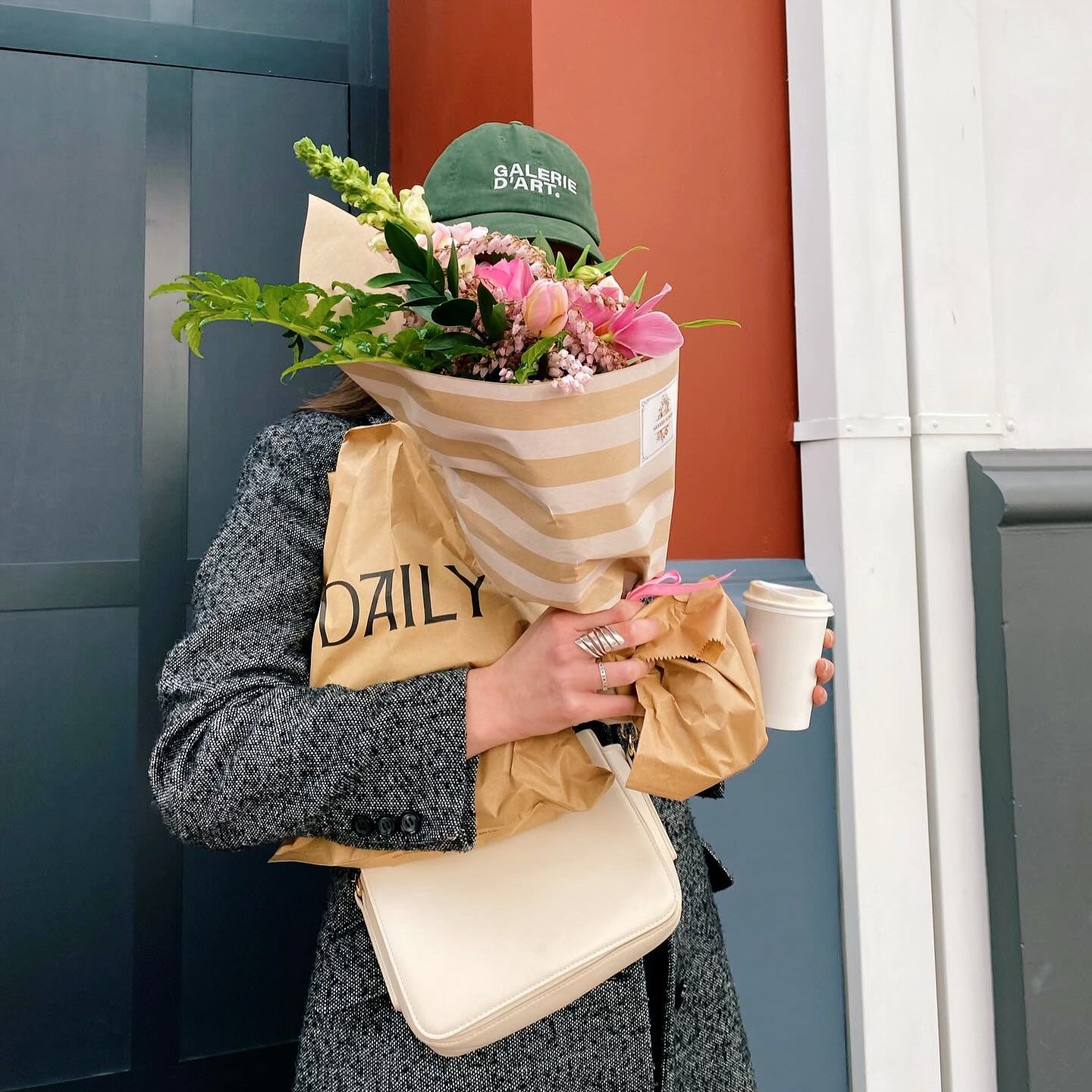 Tate stands in the middle of the frame wearing a cap and a coat. Her face is hidden by a bouquet, which she holds in her arms alongside two paper bags and a takeaway coffee cup. One of the bags says DAILY. Her handbag hangs below the other items in her hands.