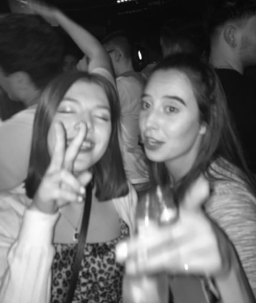A black and white image of me (white, brunette woman) with closed eyes and throwing a peace sign, dancing with my flatmate (another white, brunette woman) with crowds behind us.