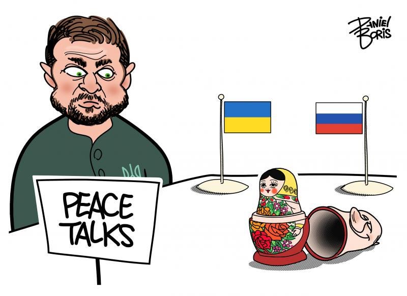 Zedlenskyy seen at left, at a tabletop that has a small Ukrainian flag placed next to him. Across from them is seen an equally small Russian flag on the table. In the foreground, on the Russian side of the table is seen a Russian nesting doll with the top of the first doll resting on its side. That piece resembles Putin's head with a smile on it.