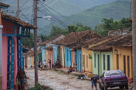 A 3x3 grid of nine photographs depicting life in Cuba. Images include: a boy diving into water; children sitting by a wall; flamingos in a lake; colorful colonial street scenes with classic cars and horse-drawn carts; a cat and a dog in a window; and a lamp-lit cobblestone street at night.