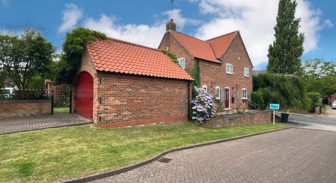 A brick house with a garage with red door and a driveway with a small lawn in front of it and trees around it