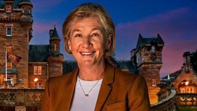 A woman in a white top and burnt organge/brown blazer smiles to camera, set against the backdrop of a castle by night A woman in a white top and burnt organge/brown blazer smiles to camera, set against the backdrop of a castle by night