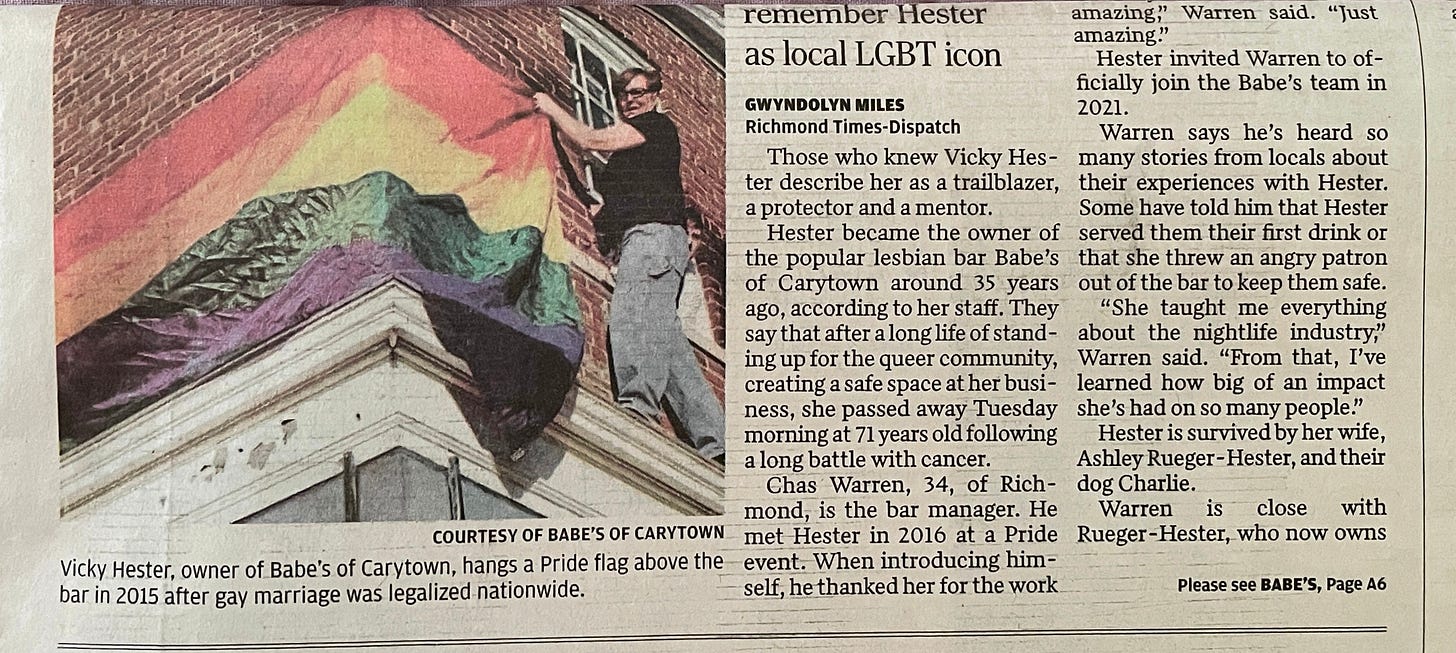 woman wearing glasses, black t-shirt, and jeans standing on a narrow ledge as she hangs a rainbow flag on a brick wall