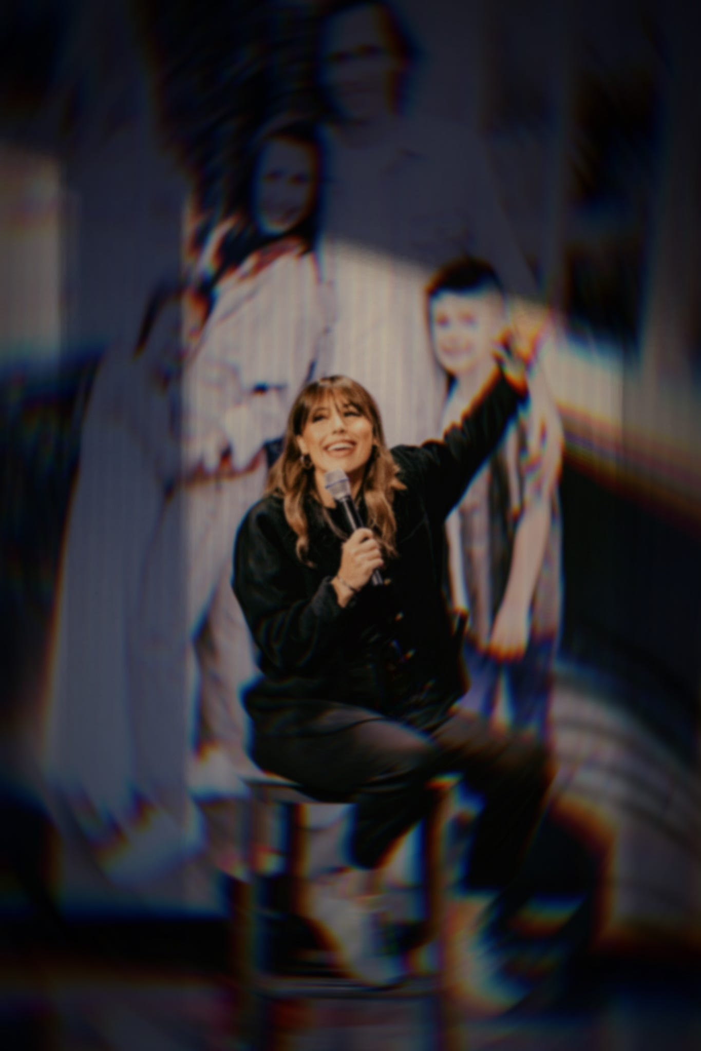 Woman holding microphone, smiling with arm raised on stage in front of blurred family image projection. Symbolic photo illustrating vulnerability, public ministry, trauma, and personal healing—featured in Substack post titled "f/2.8."