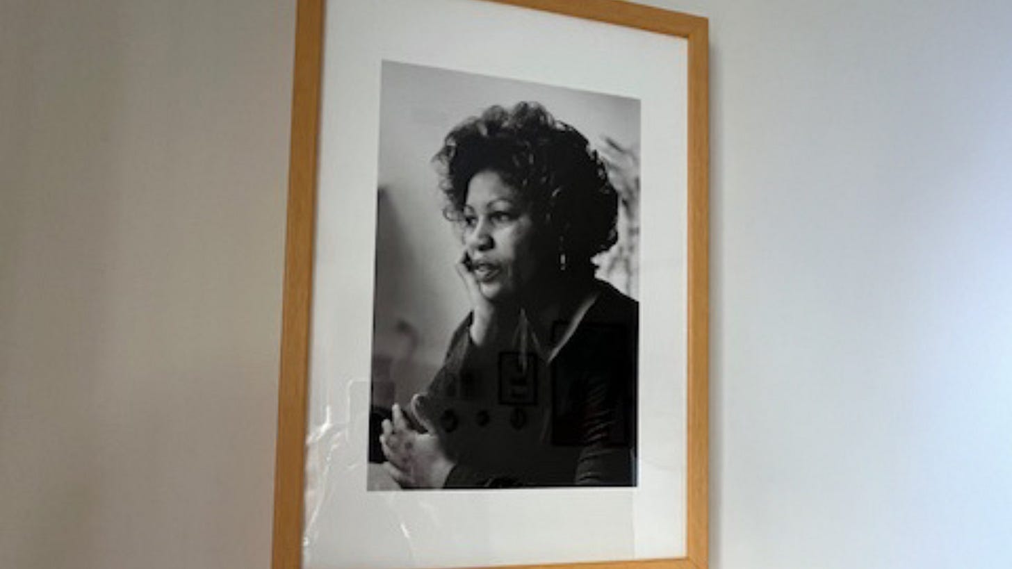Black and white portrait photograph of Toni Morrison in a wooden frame with white matting, mounted on a white wall. Morrison is shown in a contemplative pose with her hand near her face, wearing dark clothing and displaying her characteristic curly hairstyle.