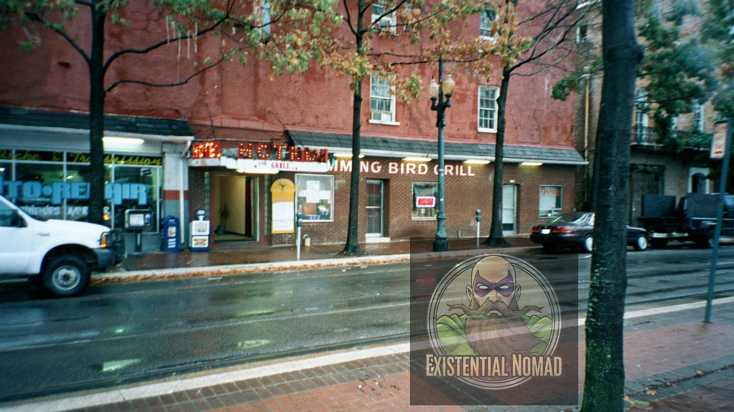  This is a photograph of a wet street in front of a brick building with several storefronts. The street is lined with bare-branched trees, and the light reflects off the rain-slicked pavement. The most prominent sign on the building reads "HUMMING BIRD GRILL" in red neon lettering. To the left, a white truck is partially visible, and to the right, a black sedan is parked behind a tree. The sky is overcast and the overall atmosphere is somewhat gloomy.