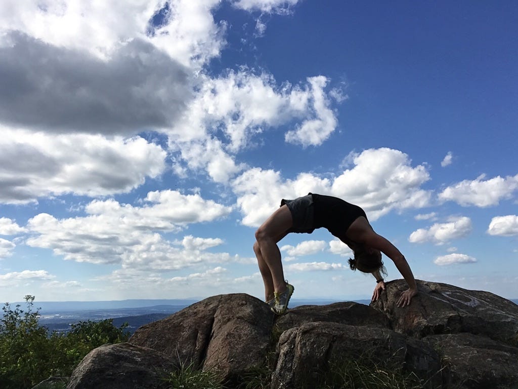 A woman does a yoga backbend on a mountaintop in upstate New York, in silhouette against a blue sky with spotty clouds.