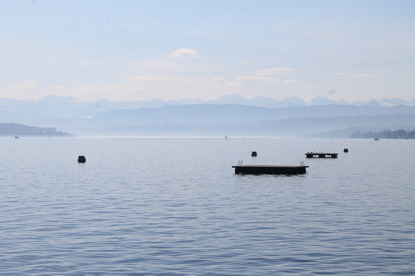 Fotografia di un lago dove l'acqua è tranquilla e su cui galleggiano alcune piccole piattaforme; in lontananza si vedono delle colline e, oltre, i profili delle montagne Fotografia di un lago dove l'acqua è tranquilla e su cui galleggiano alcune piccole piattaforme; in lontananza si vedono delle colline e, oltre, i profili delle montagne