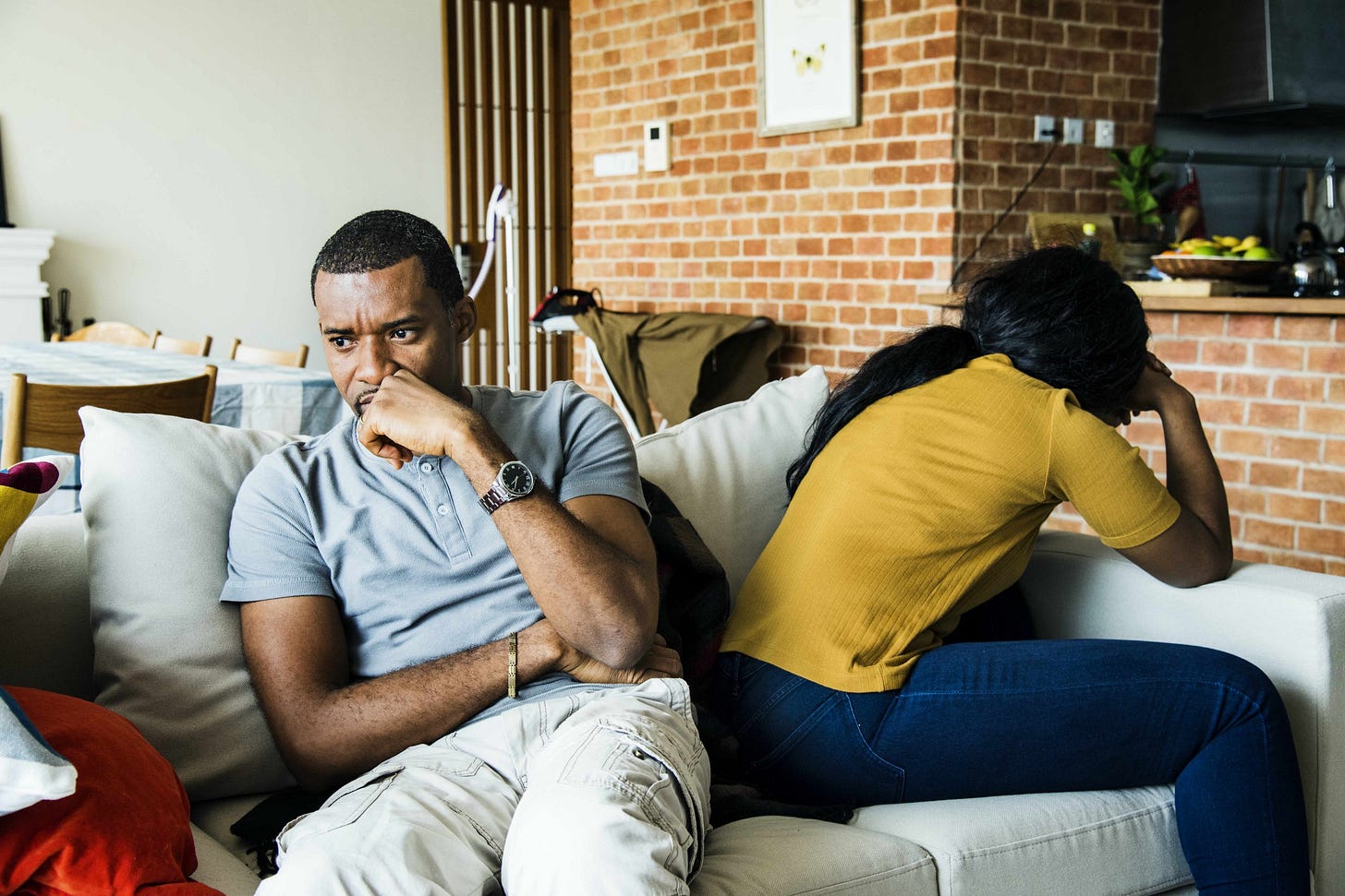 An upset couple sit on either end of the couch. Their backs face one another.