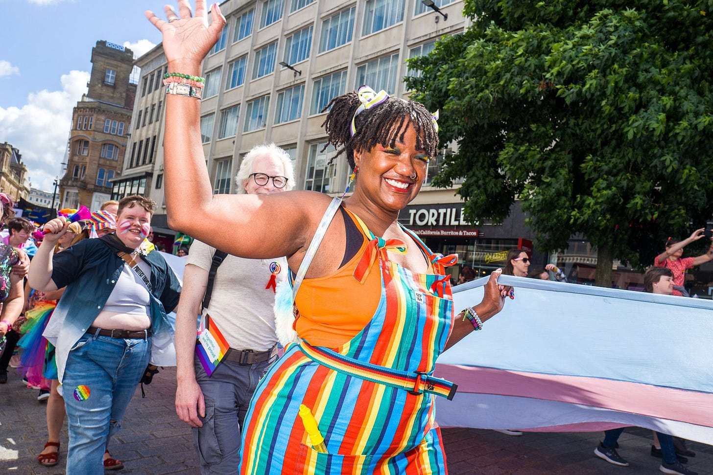 A black person waves to camera while holding part of the UK's biggest trans flag. They have a rainbow set of dungarees on.
