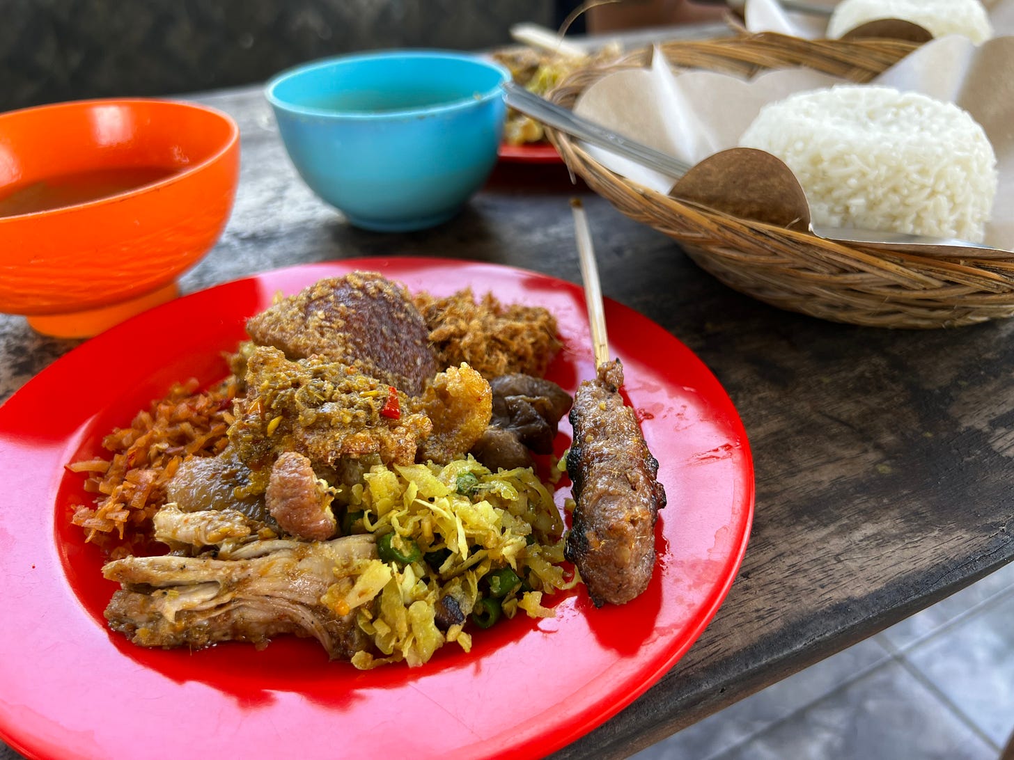 A red plastic plate of babi guling with shredded pork, long beans and coconut, a grilled minced-meat skewer and crackling, beside broth and rice.