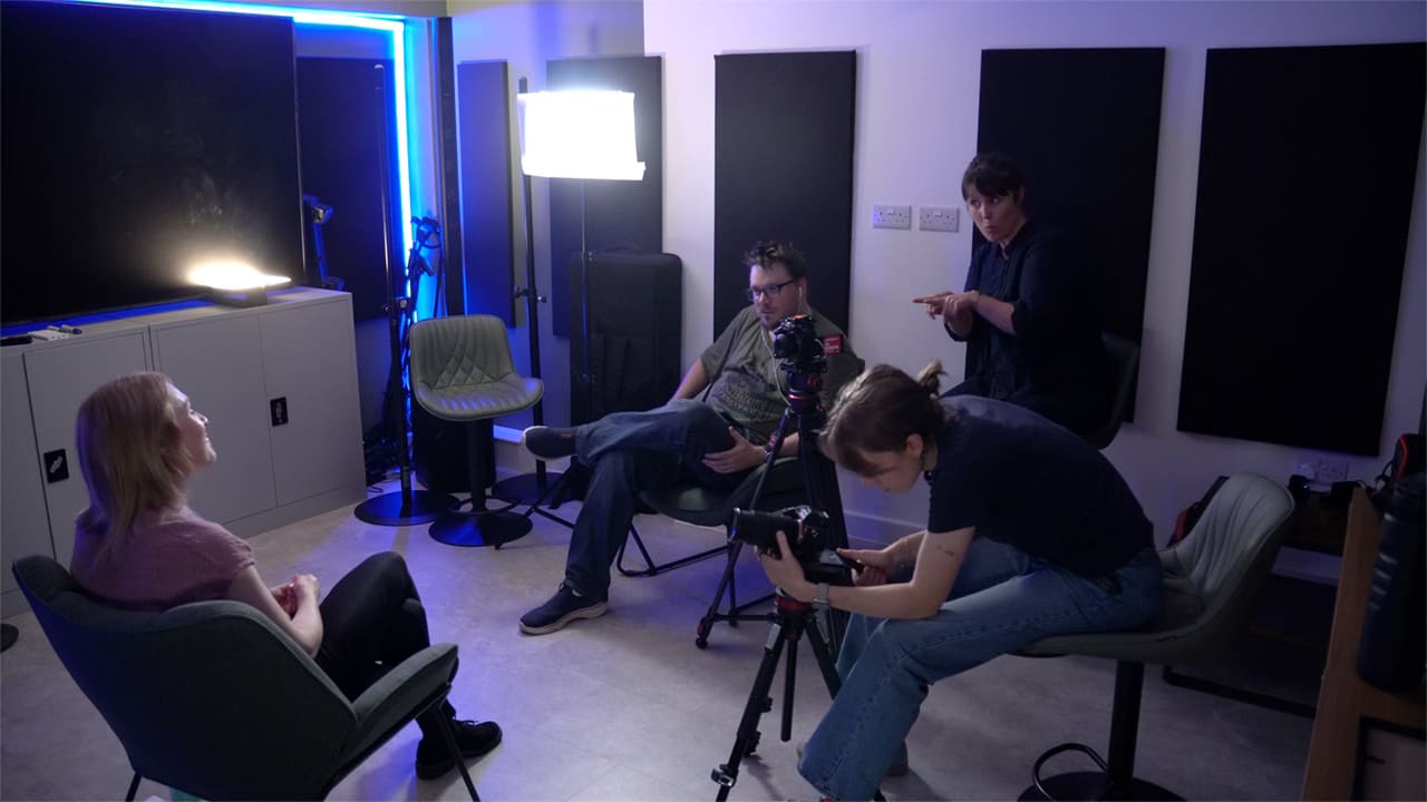 A man in glasses interviews a woman with blonde hair in front of lights and cameras. A woman operates the camera. Behind the interviewer a BSL sign language interpreter relays the question in sign language for the deaf interviewee. A man in glasses interviews a woman with blonde hair in front of lights and cameras. A woman operates the camera. Behind the interviewer a BSL sign language interpreter relays the question in sign language for the deaf interviewee.