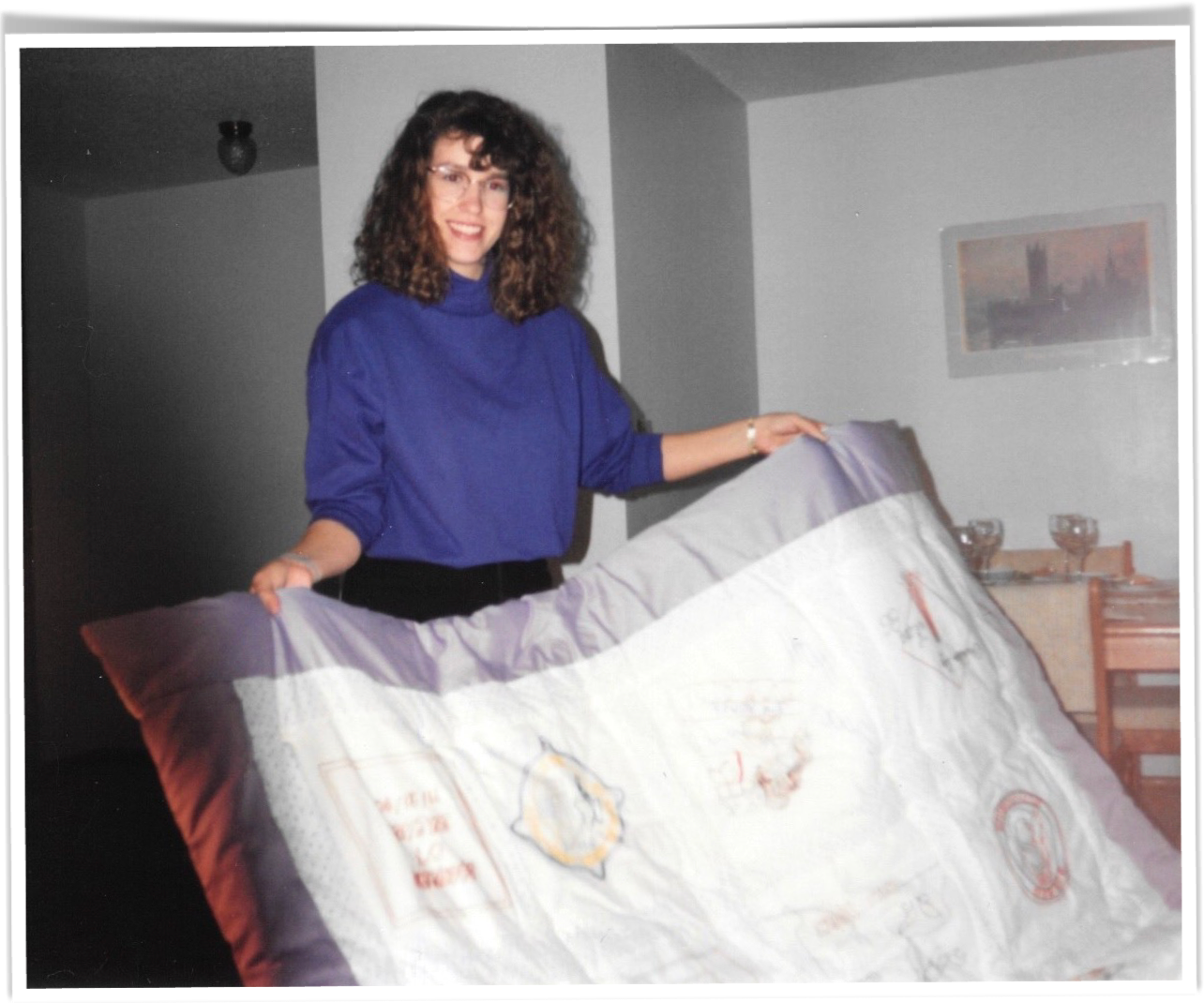 Woman smiling, wearing glasses and holding a large hand-made quilt with embroidered blocks representing moments of her life.