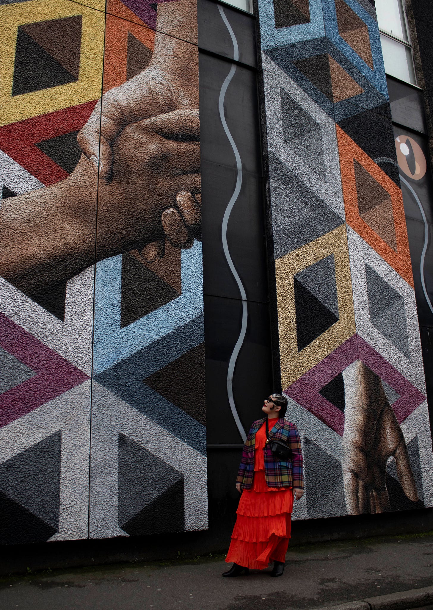 Jennifer Heinen in a bright orange tiered dress and tartan blazer, looking up at a large geometric street mural with two oversized hands shaking.