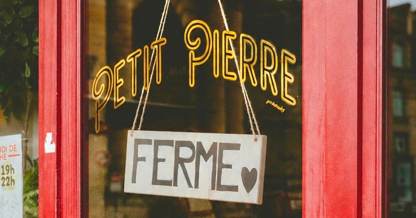 A shop window with "Petit Pierre" hand-lettered in gold on the glass, framed in red-painted wood. A small white wooden sign hangs from twine across it reading "FERMÉ" in black capitals, with a hand-drawn heart. A partly visible hours sign sits in the lower left. A shop window with "Petit Pierre" hand-lettered in gold on the glass, framed in red-painted wood. A small white wooden sign hangs from twine across it reading "FERMÉ" in black capitals, with a hand-drawn heart. A partly visible hours sign sits in the lower left.