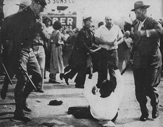 Black man on ground, surrounded by white police carrying sticks