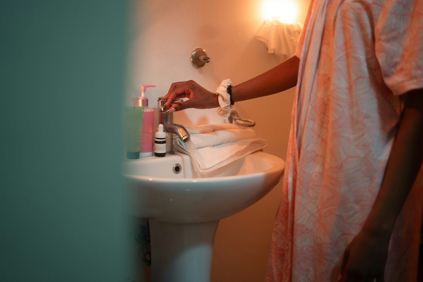 A person in a floral robe reaches toward a bathroom sink surrounded by skincare products in warm lamplight, capturing the intimate private ritual that precedes the day, the kind of specific personal sequence that belongs to the authentic creative process but rarely gets described.