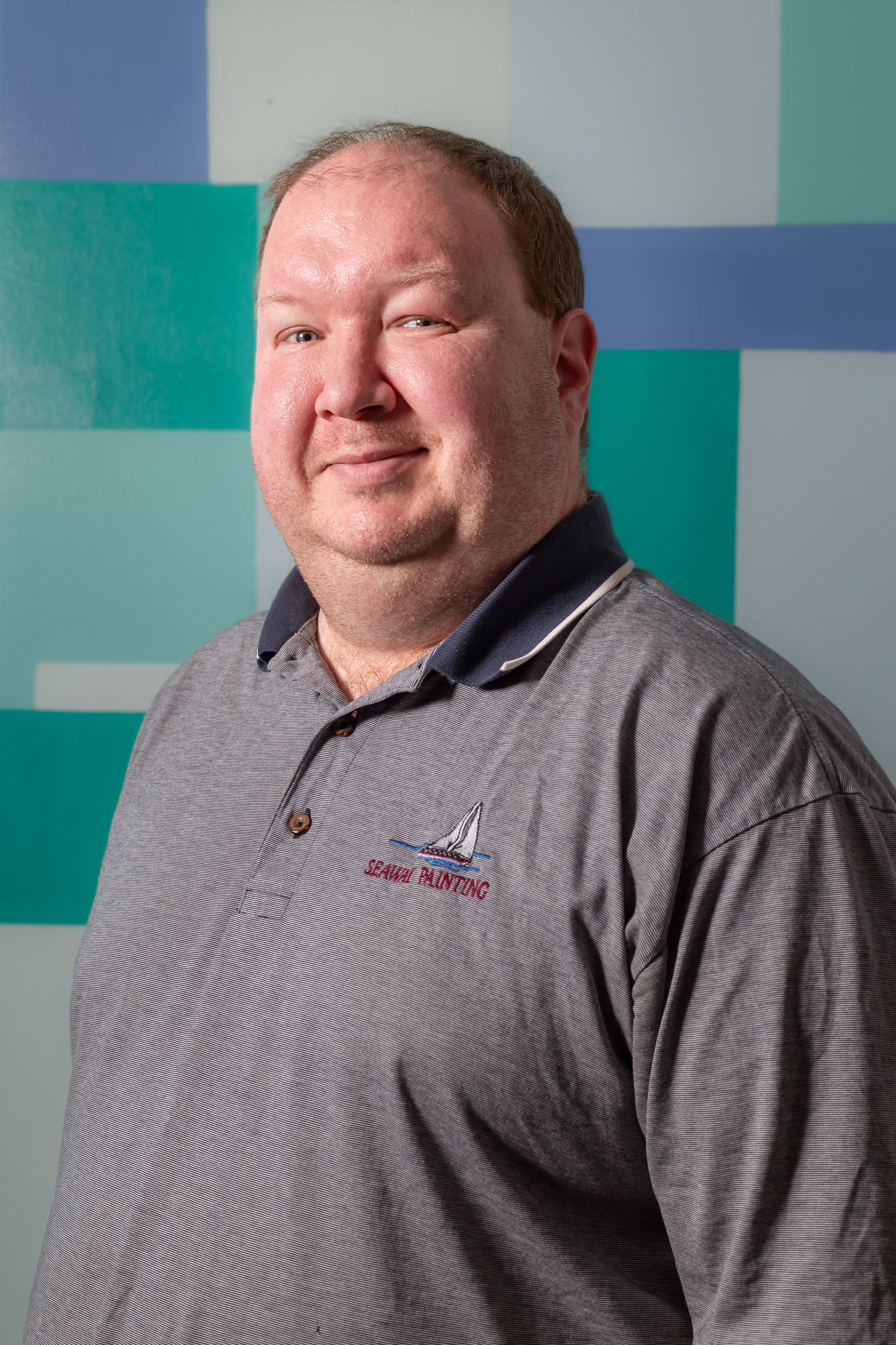 Portrait of a 42 year old white skinned man with light green eyes looks at the camera with a slight smile. He has thinning, short, brown hair. He wears a gray polo shirt with a navy blue collar.