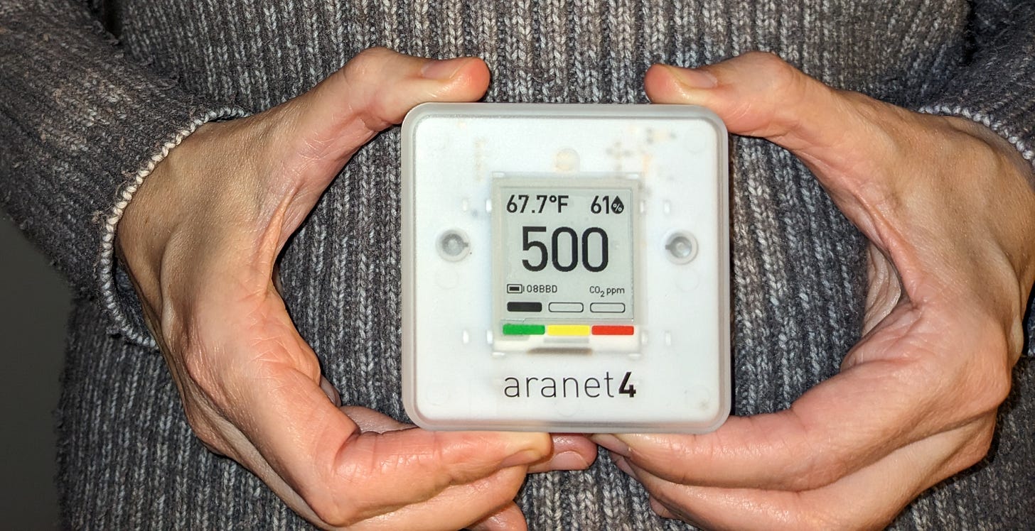 Cropped photo of me: all you can see is my grey knit sweater and my hands holding an Aranet4. It's a white plastic box 7cm by 7cm by 2cm with a display that reads: "67.7℉, 61% humid, 500ppm CO2." 