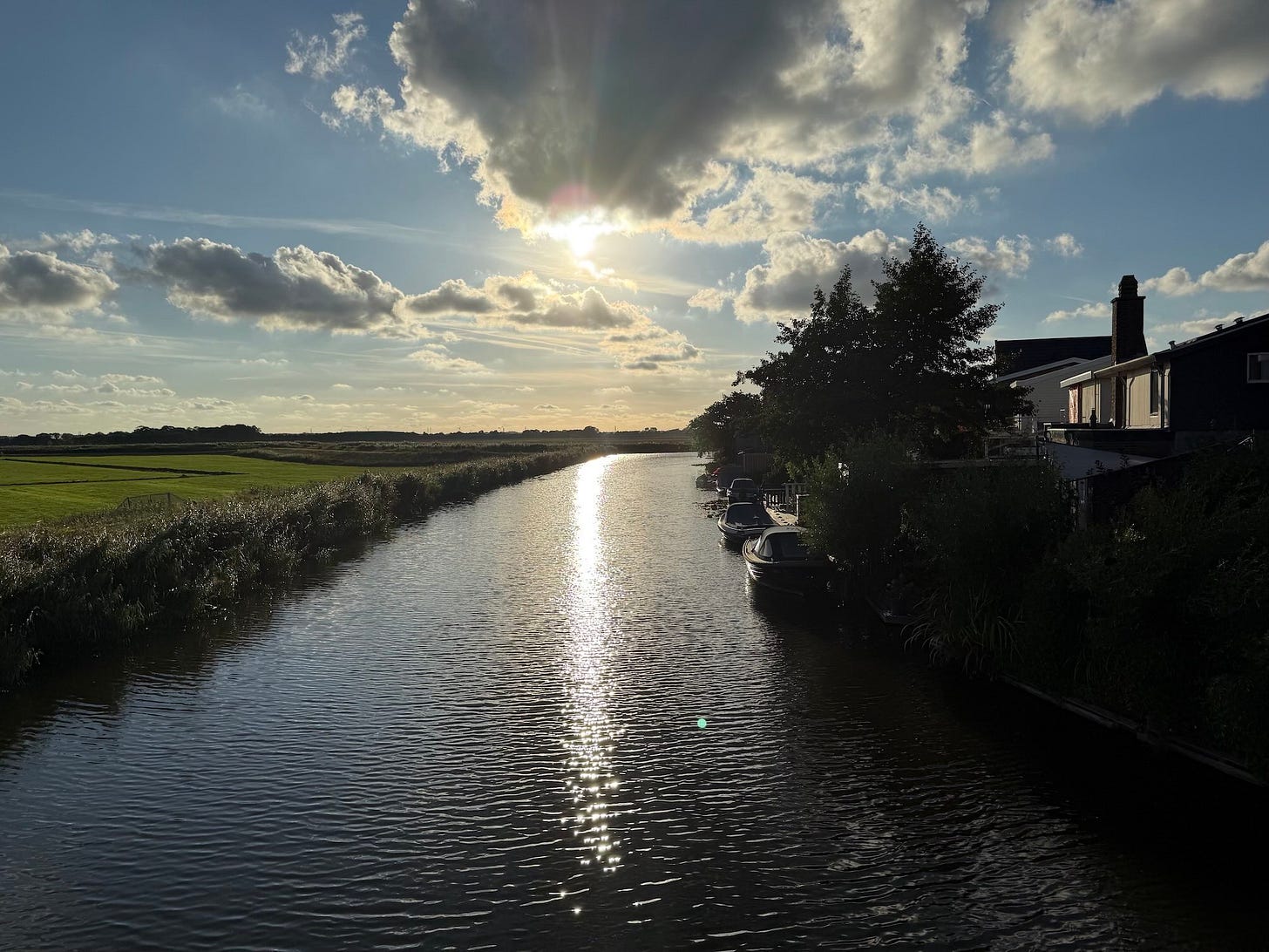 A peaceful canal in North Holland at sunset, with the sun low in the sky casting golden reflections on the water. Boats are moored along the right bank near houses and trees, while open green fields stretch out to the left under scattered clouds.