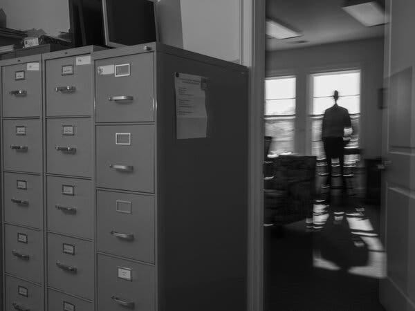 A man is seeing blurred in the background as he stands in front of a window in an office. The foreground of the photo is a collection of filing cabinets. A man is seeing blurred in the background as he stands in front of a window in an office. The foreground of the photo is a collection of filing cabinets.