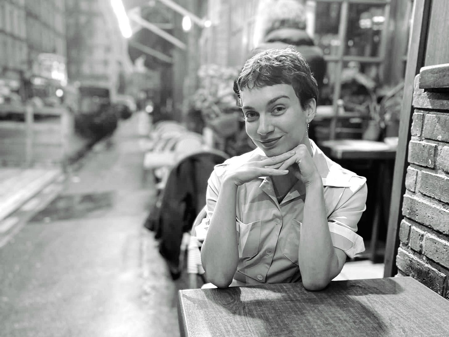 A black and white photo of Leonie, streetside at a wooden table, with her chin resting on her hands and her elbows resting on the table. A sweet smile and kind eyes bless her face. 