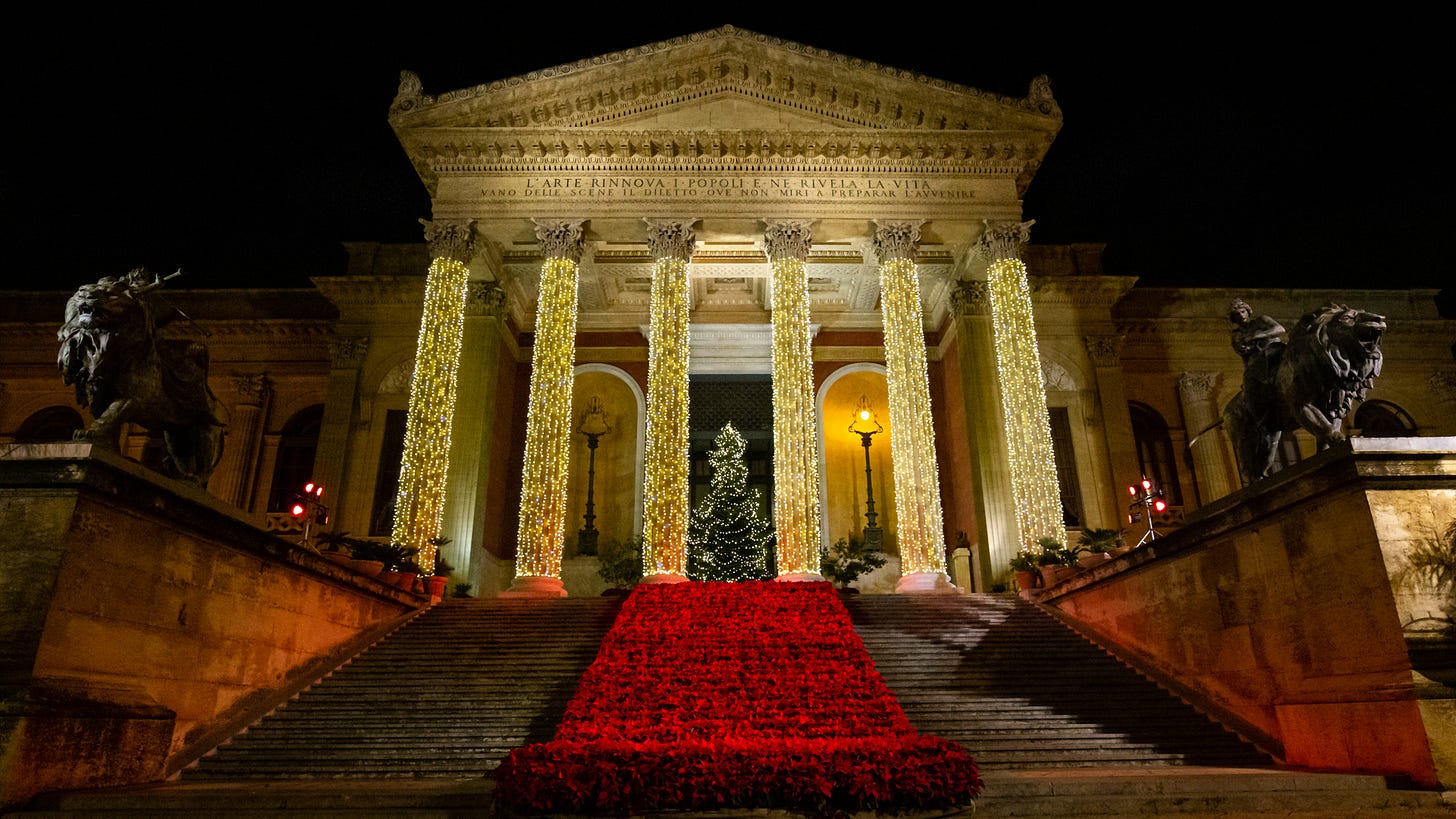 The portico and steps of an ornate building are decorated for Christmas with lights, Christmas trees and a red carpet.