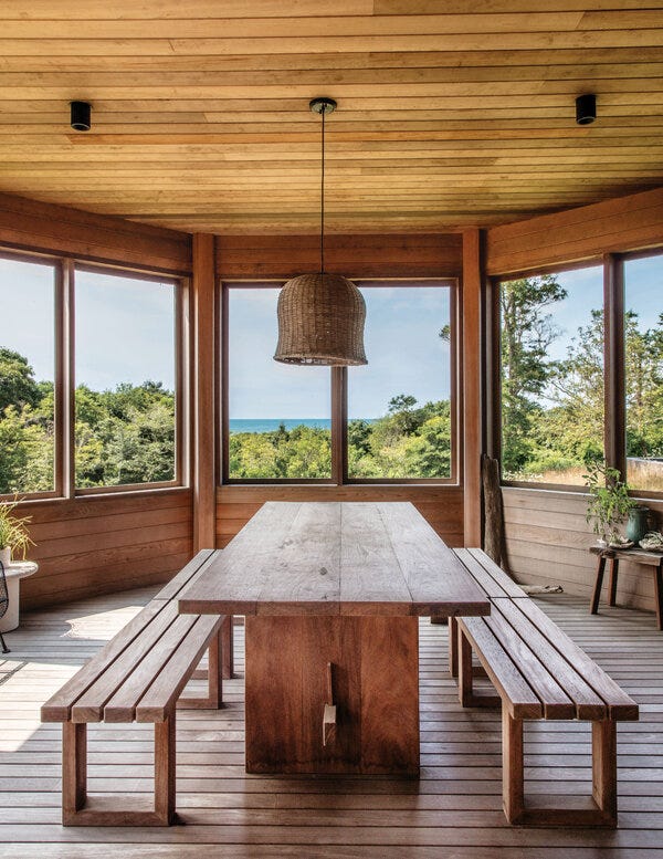 A wooden porch, with windows through which trees and a sliver of ocean are visible. In the center, a long wooden table with two benches on either side.