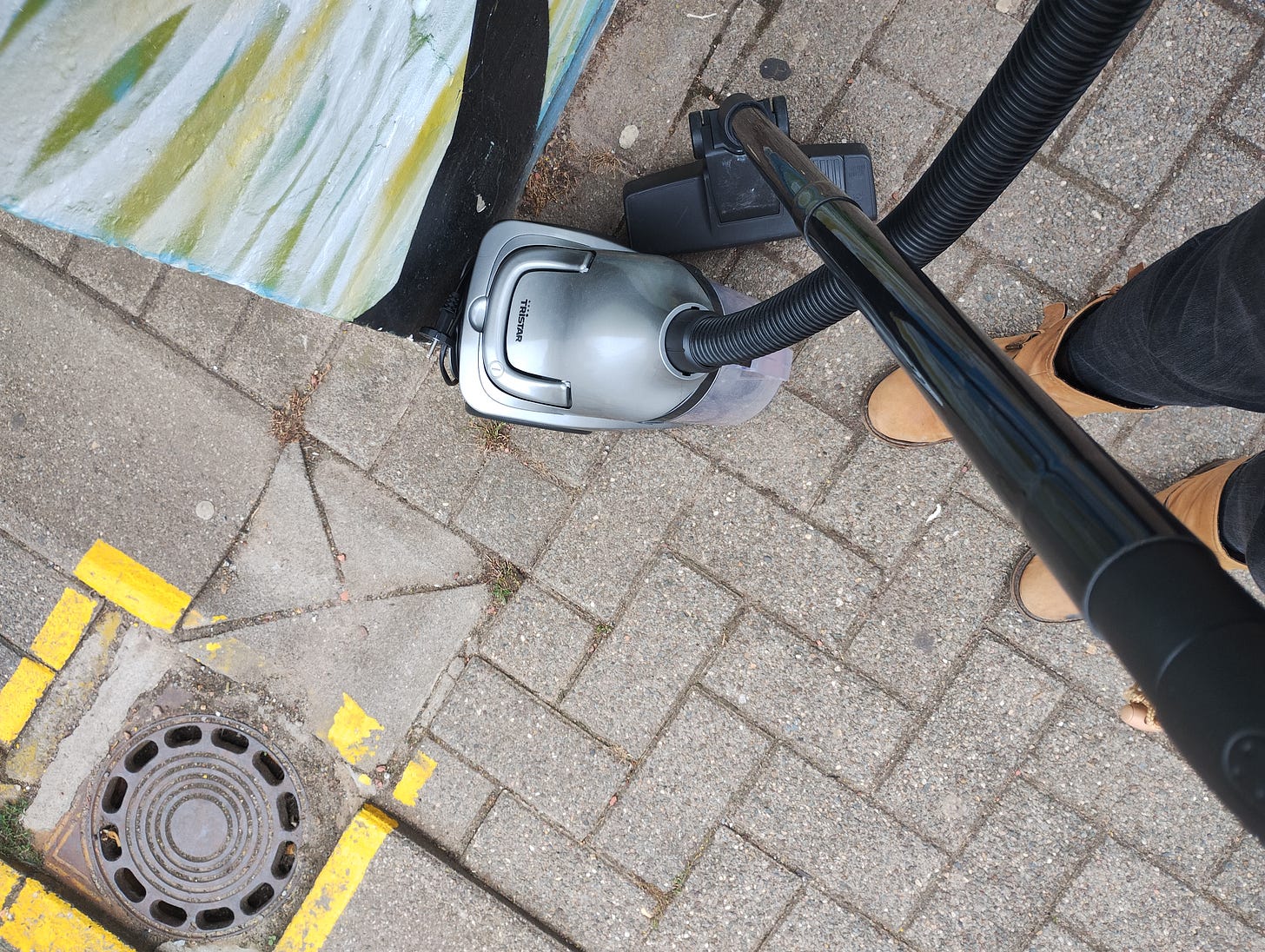 Picture of two legs in black jeans and brown boots next to a vaccuum cleaner on a grey pavement
