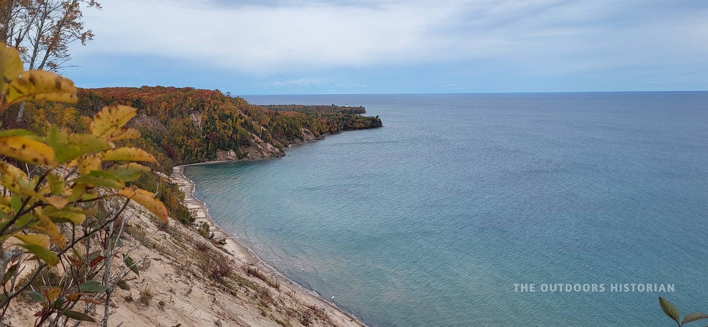 Autumn coastline of Pictured Rocks National Lakeshore. Towering sand dunes in the foreground descend toward a low, wooded point where the white Au Sable Lighthouse stands in the distance. The shoreline is bordered by a thick canopy of red and gold fall trees, contrasting with the pale sand and the blue of the lake.